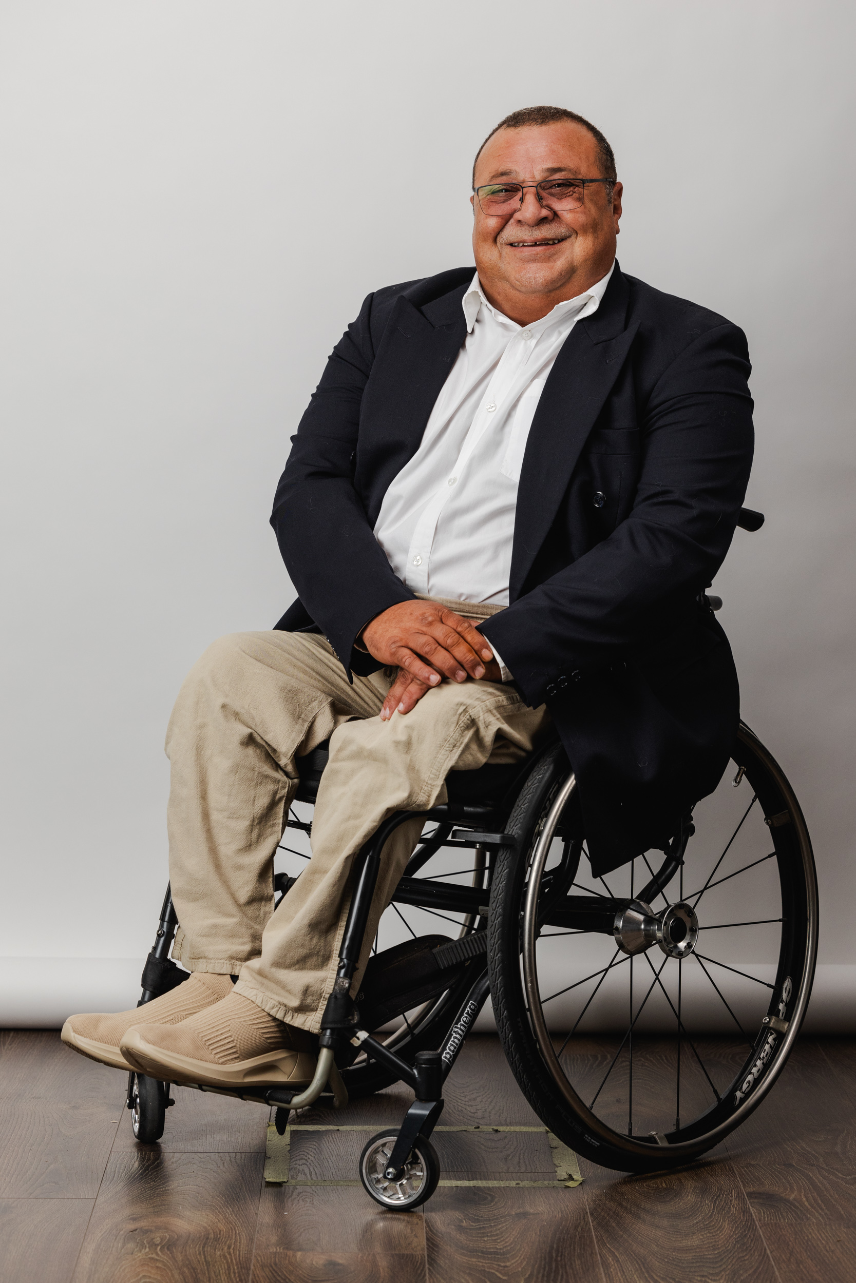Portrait d'un homme en fauteuil roulant, souriant, vêtu d'un blazer et d'une chemise blanche, assis dans un studio avec un fond neutre.